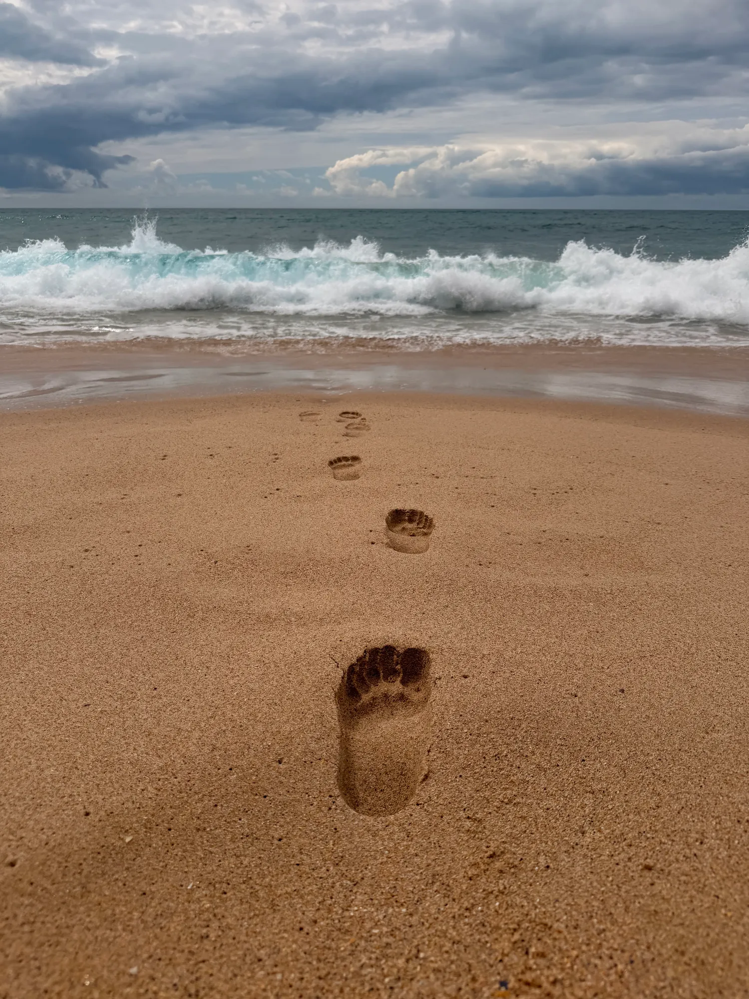 Bare footprints trailing across golden sand toward the Atlantic surf under a cloudy sky