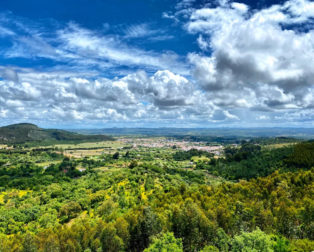 Panoramic view across the lush green Alentejo valley with a village nestled among hills under a dramatic cloud-filled sky