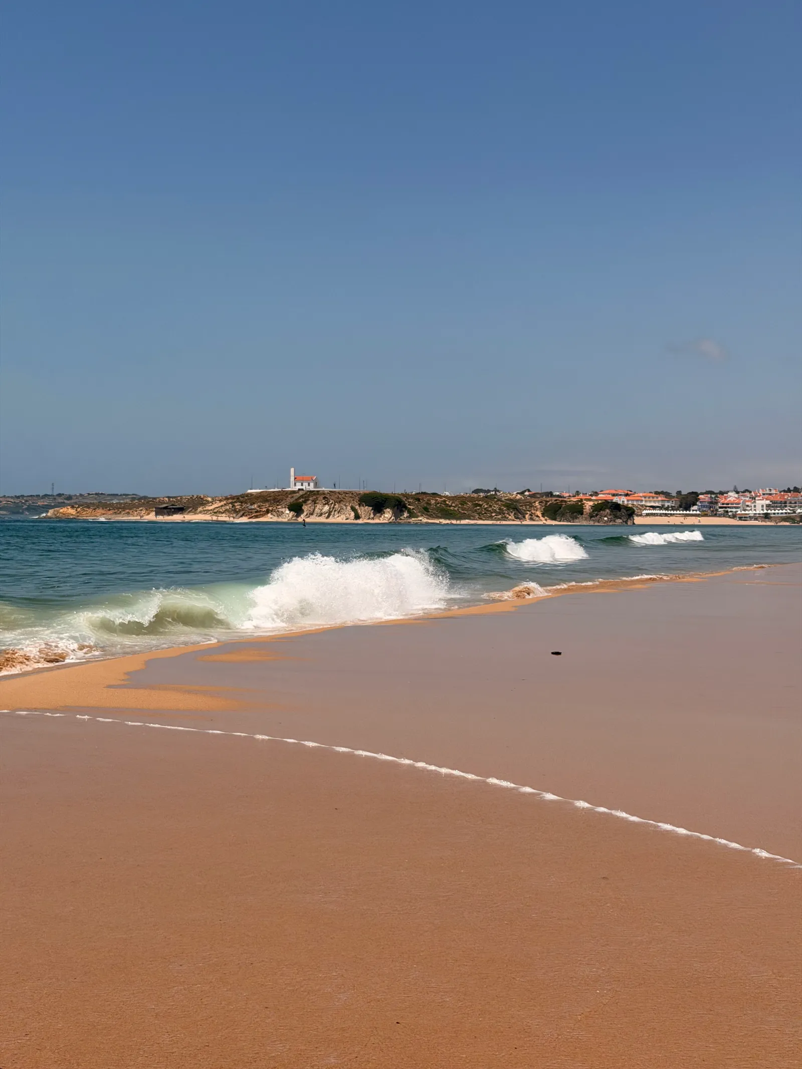 Elevated view of a long curving sandy beach with white Atlantic waves rolling in beside a coastal town under clear blue sky