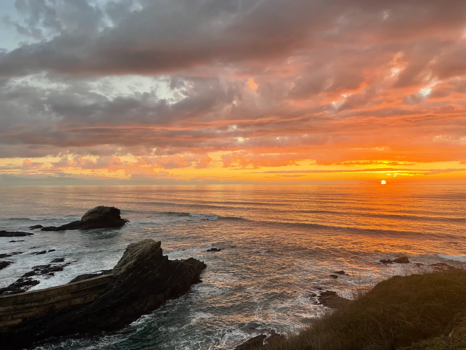 Dramatic sunset over the Atlantic with fiery orange clouds, dark rocky cliffs and white waves crashing below