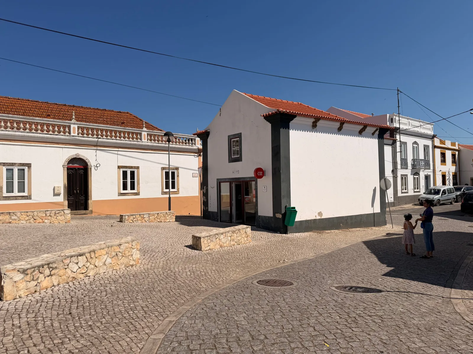Sunlit cobblestone square in São Luís lined with whitewashed houses trimmed in ochre and blue, terracotta rooftops and a small post office under a clear blue sky