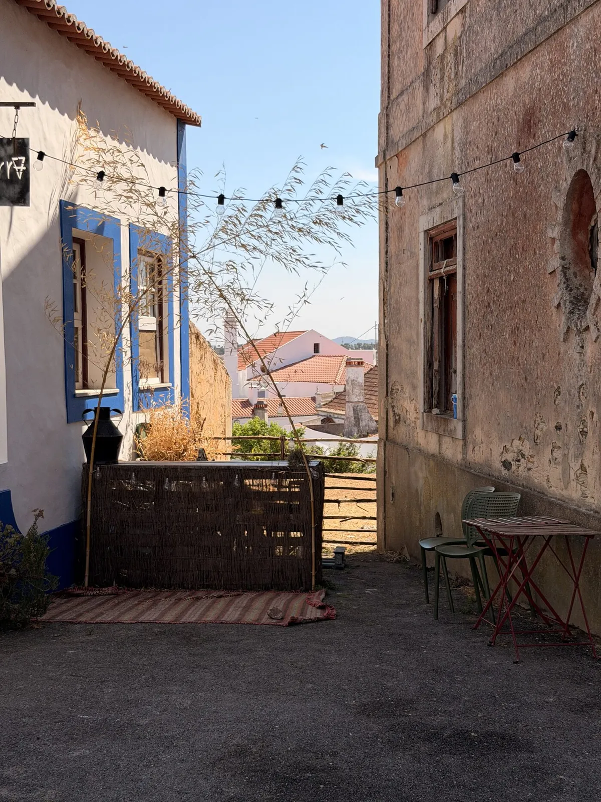 Rustic shaded terrace strung with festoon lights, a reed screen and bistro chairs, looking out over São Luís rooftops in the warm afternoon sun