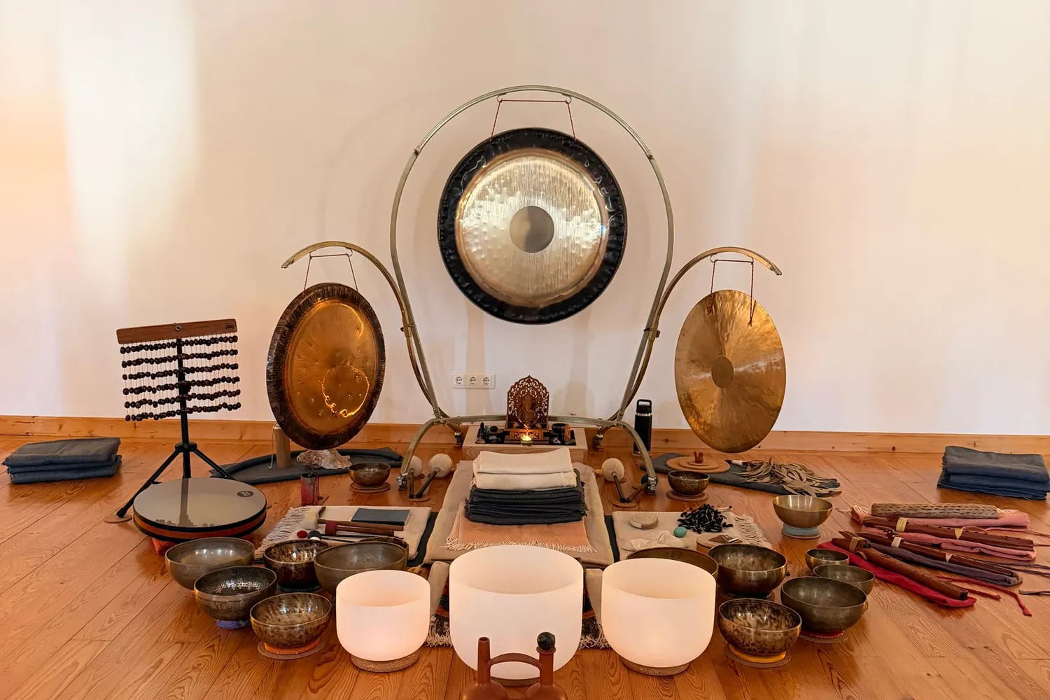 A sound healing room set up on a warm wooden floor: a large gong suspended in a brass stand between two smaller gongs, a chime tree and an altar behind, with singing bowls, crystal bowls and flutes arranged in a semicircle in front of blankets laid out for the participants
