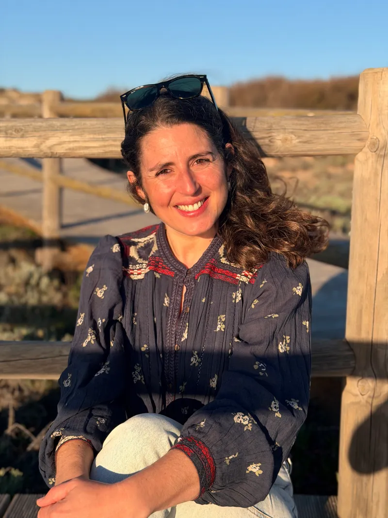 Ana smiling in warm late-day sun, sitting against a weathered wooden boardwalk railing among the coastal dunes near Casa Alma