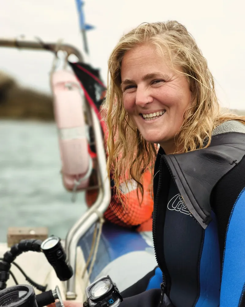 Larissa smiling in a blue and black wetsuit on a small boat, her hair wet from the water, diving gear and a cylinder stowed beside her with the estuary behind