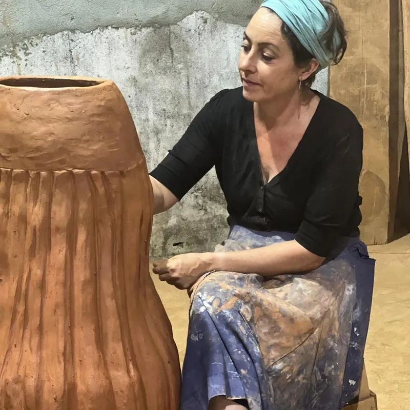 Maya seated in her studio in a clay-marked apron and turquoise headscarf, shaping the ribbed surface of a large terracotta vessel beside her