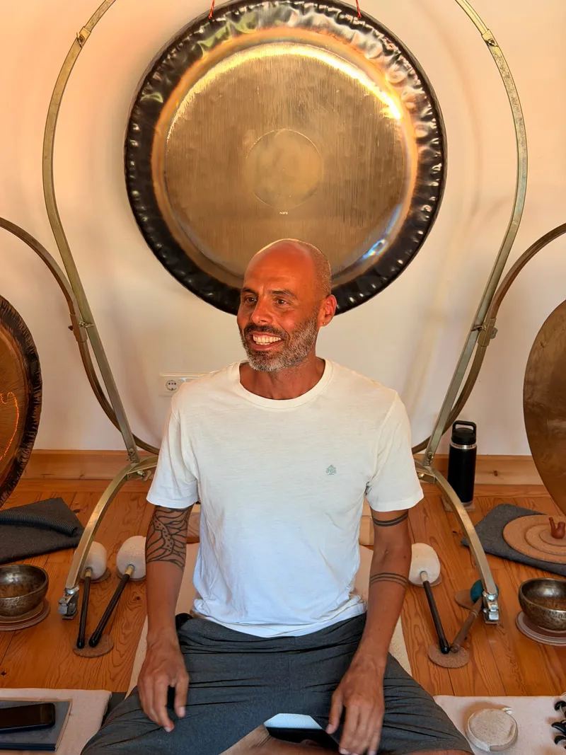 Tiago sitting cross-legged on the studio floor in a white t-shirt, smiling, with a large brass gong on its stand behind him and singing bowls and mallets laid out around him