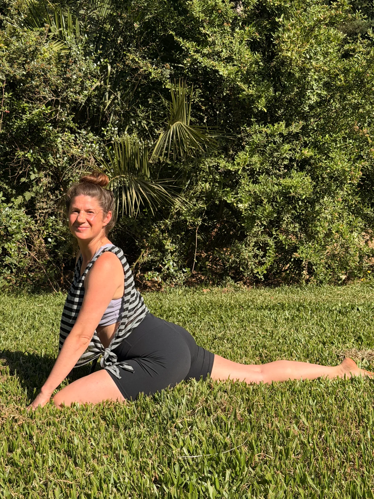 A woman holding a balancing yoga pose on sunlit garden grass beside a tall green hedge